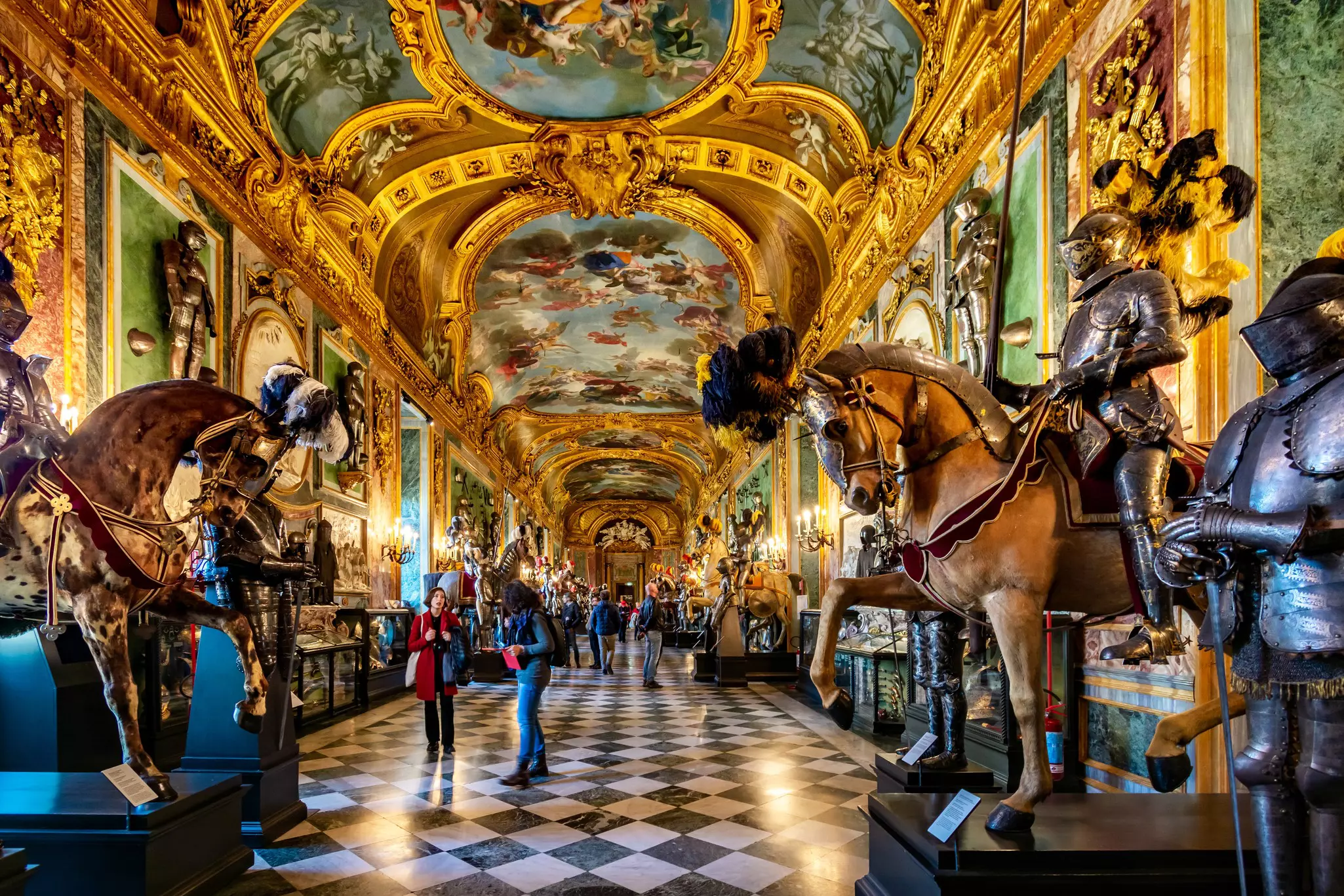 People stand on a black-and-white checked floor in a long gallery with elaborately painted ceilings, gold accents and models of armored figures on horseback.