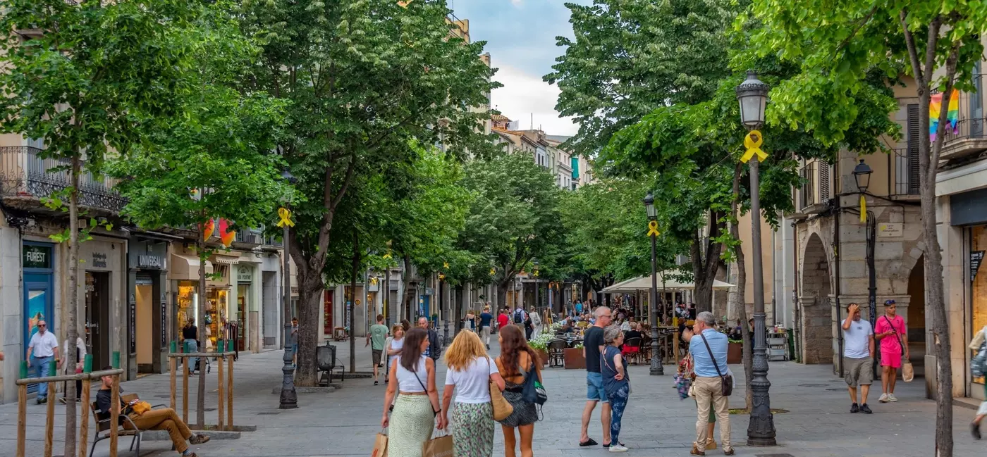 People sit at benches, dine at outdoor terraces and walk down a pedestrianized, commercial street in Spain