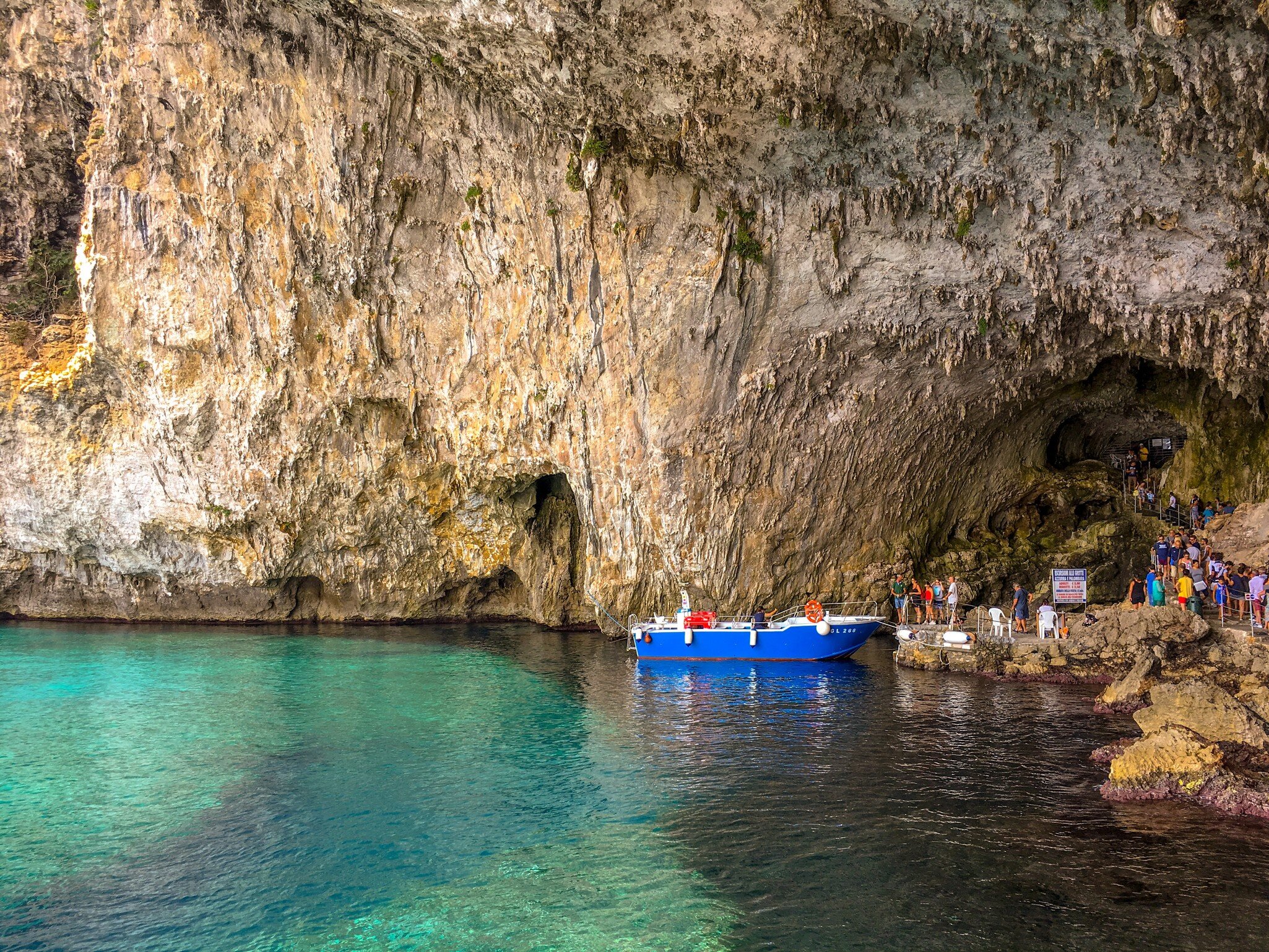 Tall streaked cliff and cave arch over clear green-blue water, with a blue boat and a line of visitors at the rocky landing