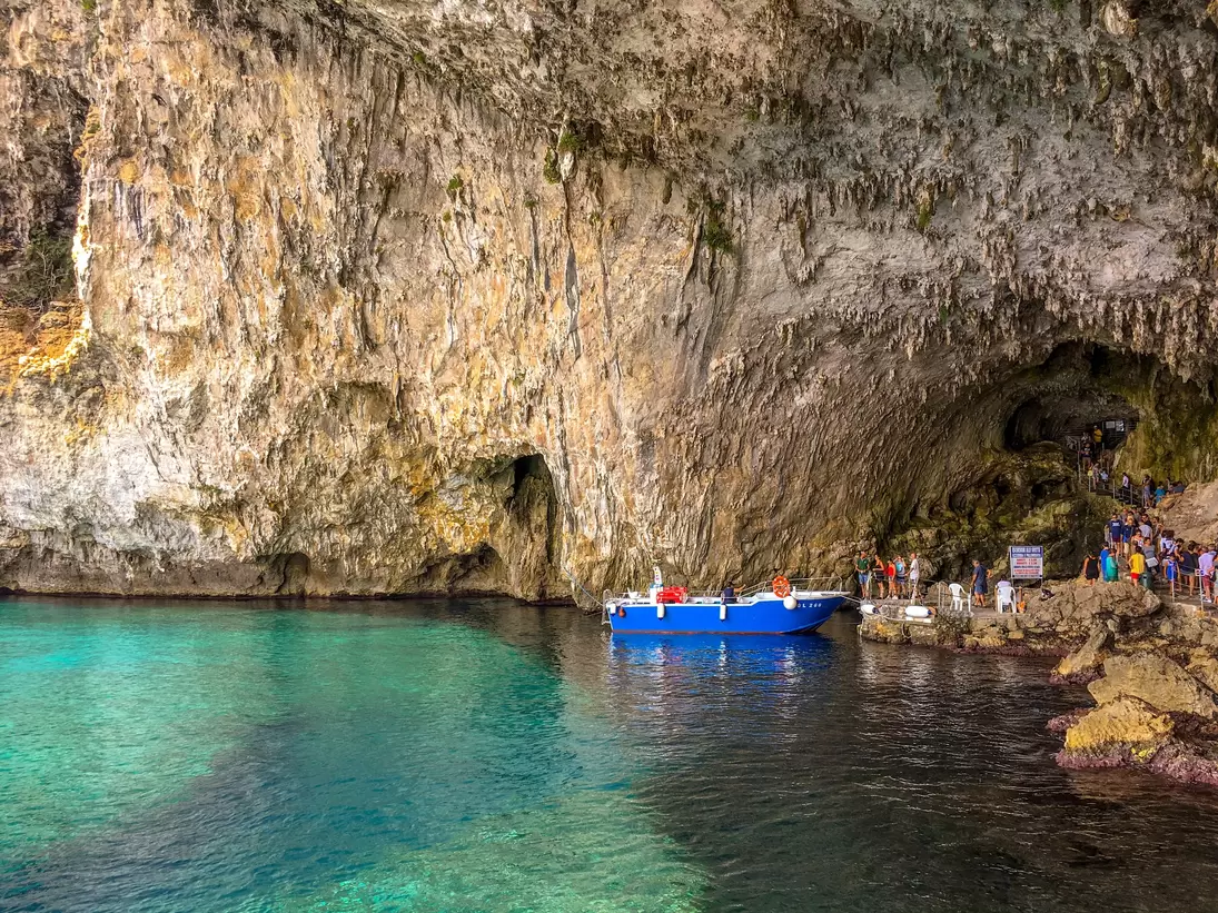 Tall streaked cliff and cave arch over clear green-blue water, with a blue boat and a line of visitors at the rocky landing