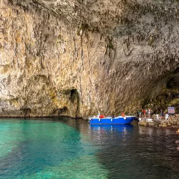 Tall streaked cliff and cave arch over clear green-blue water, with a blue boat and a line of visitors at the rocky landing