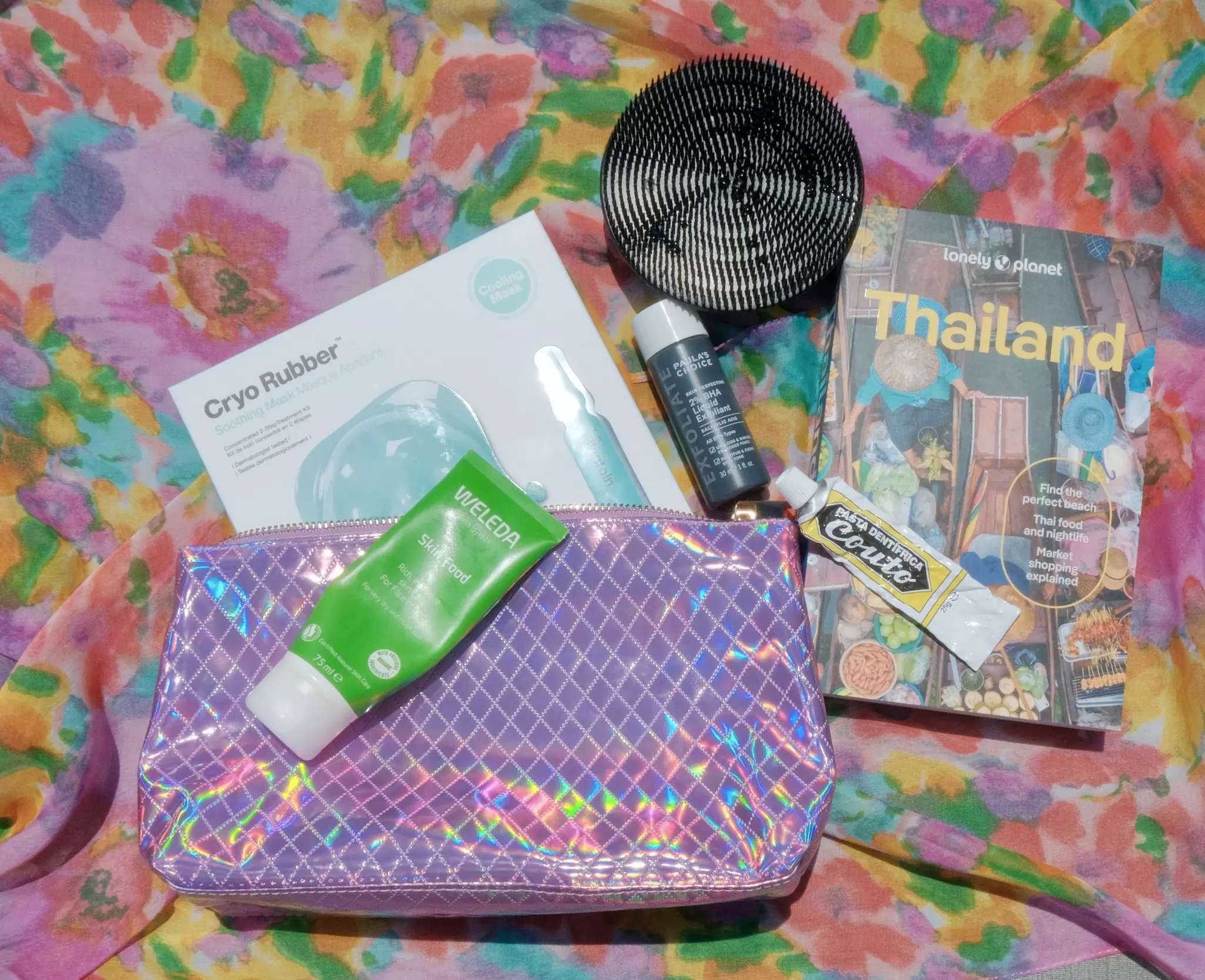 A pink metallic makeup bag, beauty products and a Thailand guidebook resting on a floral cloth.
