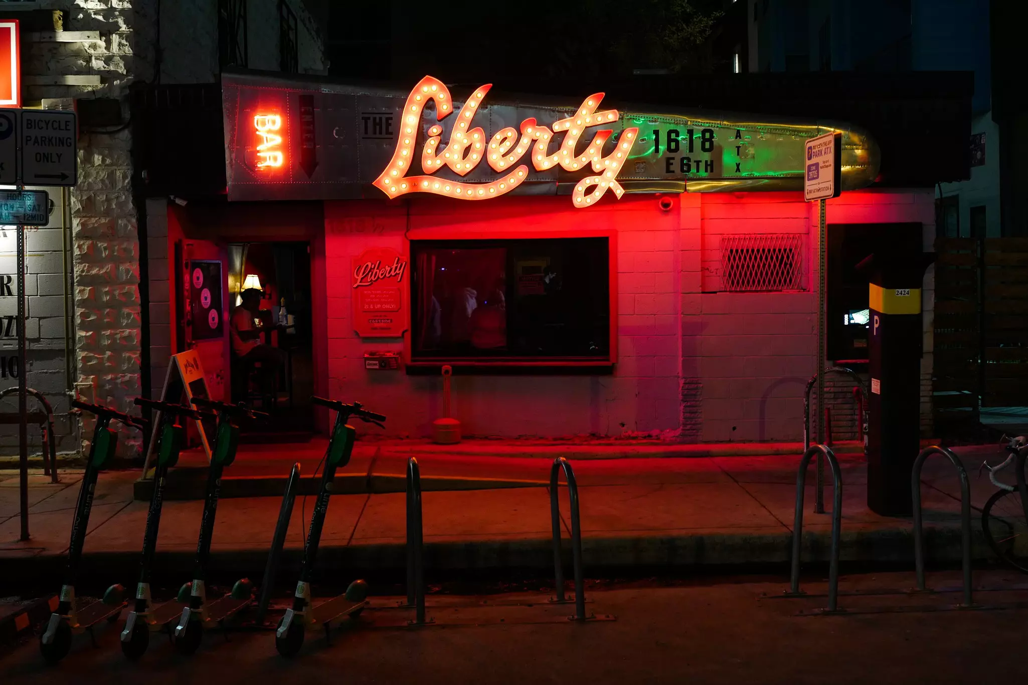The facade of a dive bar on a city street is illuminated by red lights and light-bulb letters.