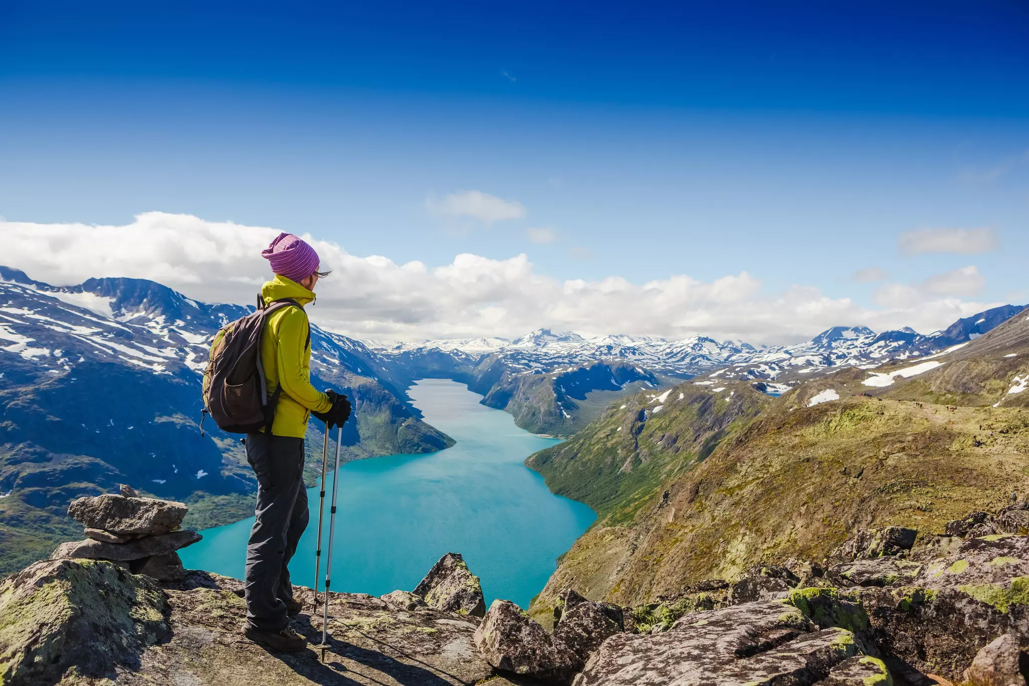 A hiker stands on a ridge in Norway looking at a blue lake and snow-covered mountains in the distance.