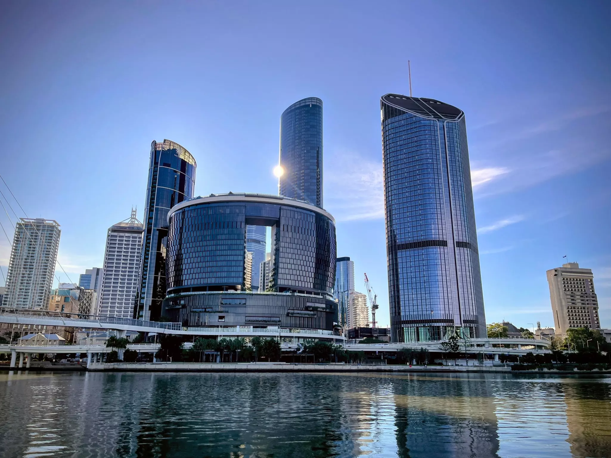 Waterfront view of a modern city skyline featuring curved glass skyscrapers reflecting sunlight, seen from across a river.