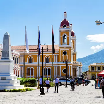 People walk past a monument in a square, with a large, yellow-painted church in the background, Granada, Nicaragua
