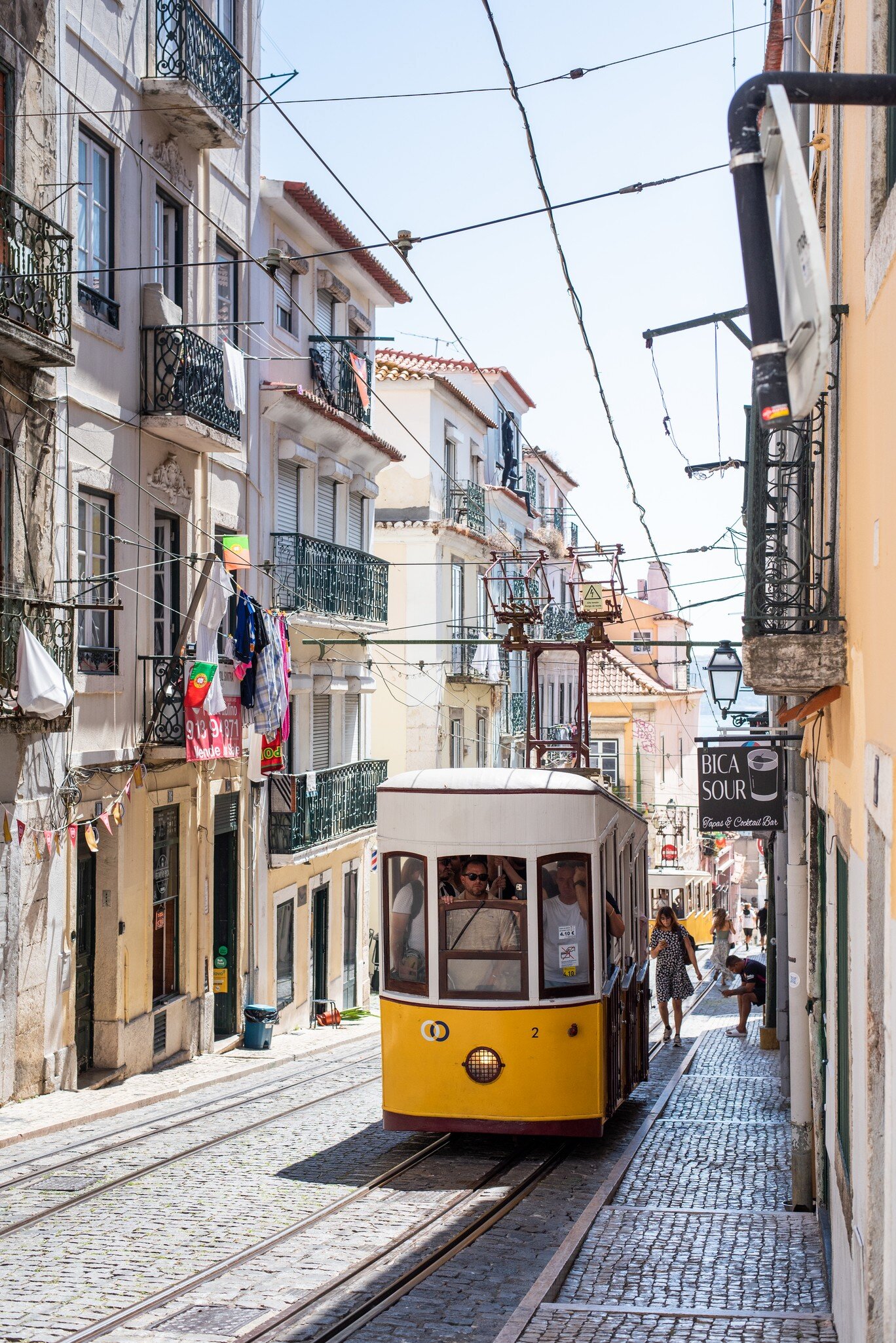 Yellow funicular car on steep cobbled tracks between balconied buildings with hanging laundry in Lisbon, Portugal.