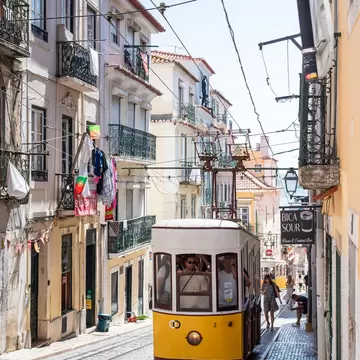 Yellow funicular car on steep cobbled tracks between balconied buildings with hanging laundry in Lisbon, Portugal.