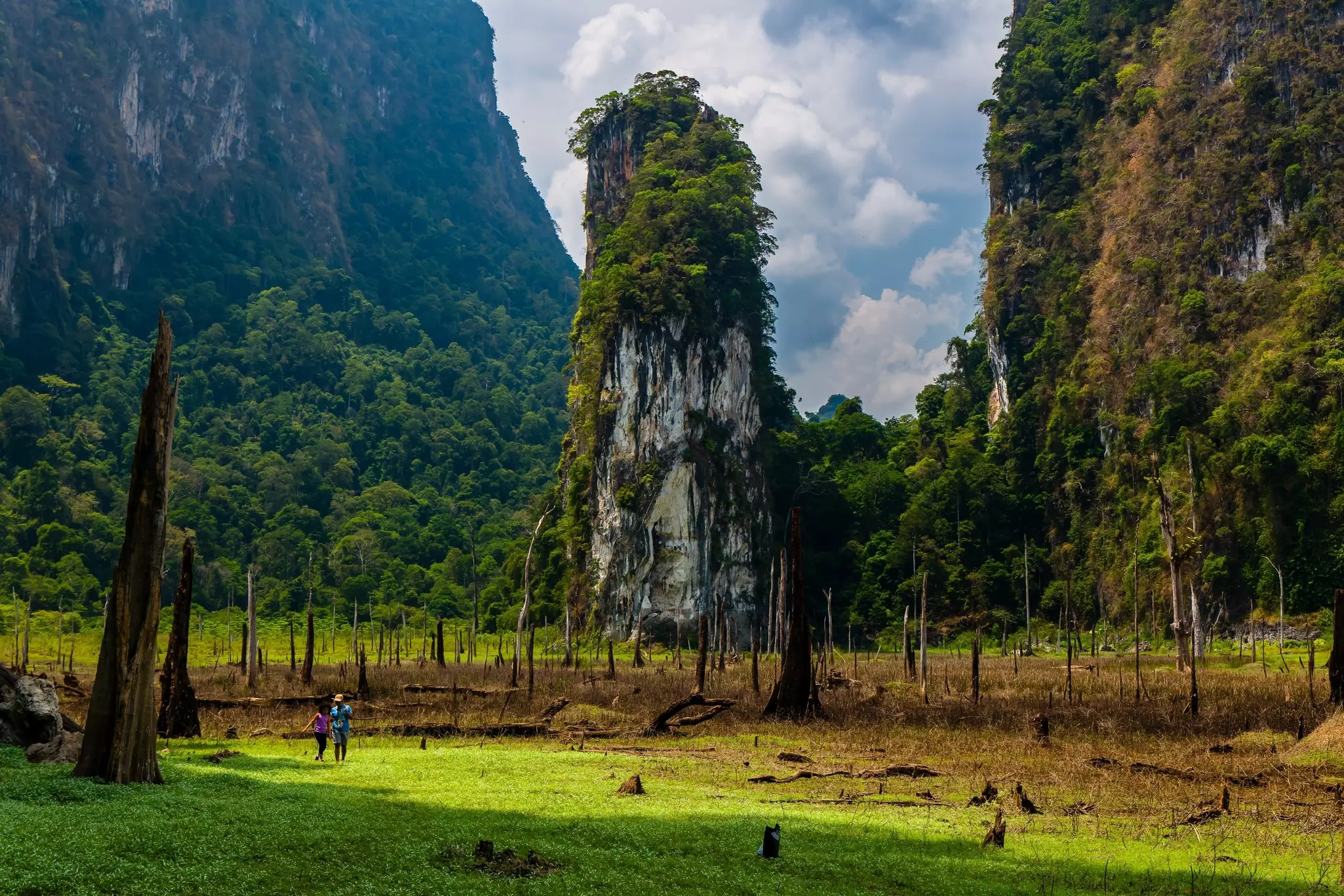 Two hikers in Thailand cross a grassy area in front of a tall karst column, with larger, vegetation-covered rocky mountains rising on either side.