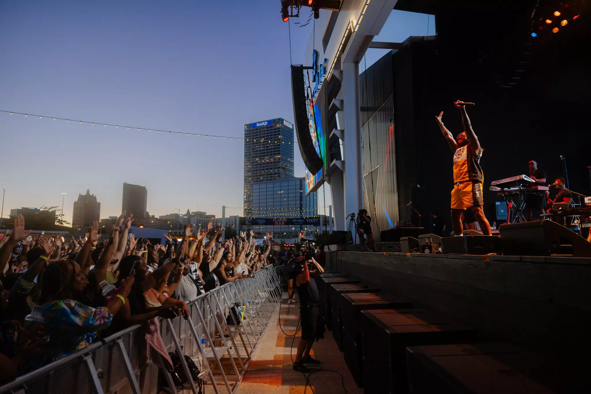 A performer takes the stage in front of a huge crowd in the evening at a bandshell in a city park.