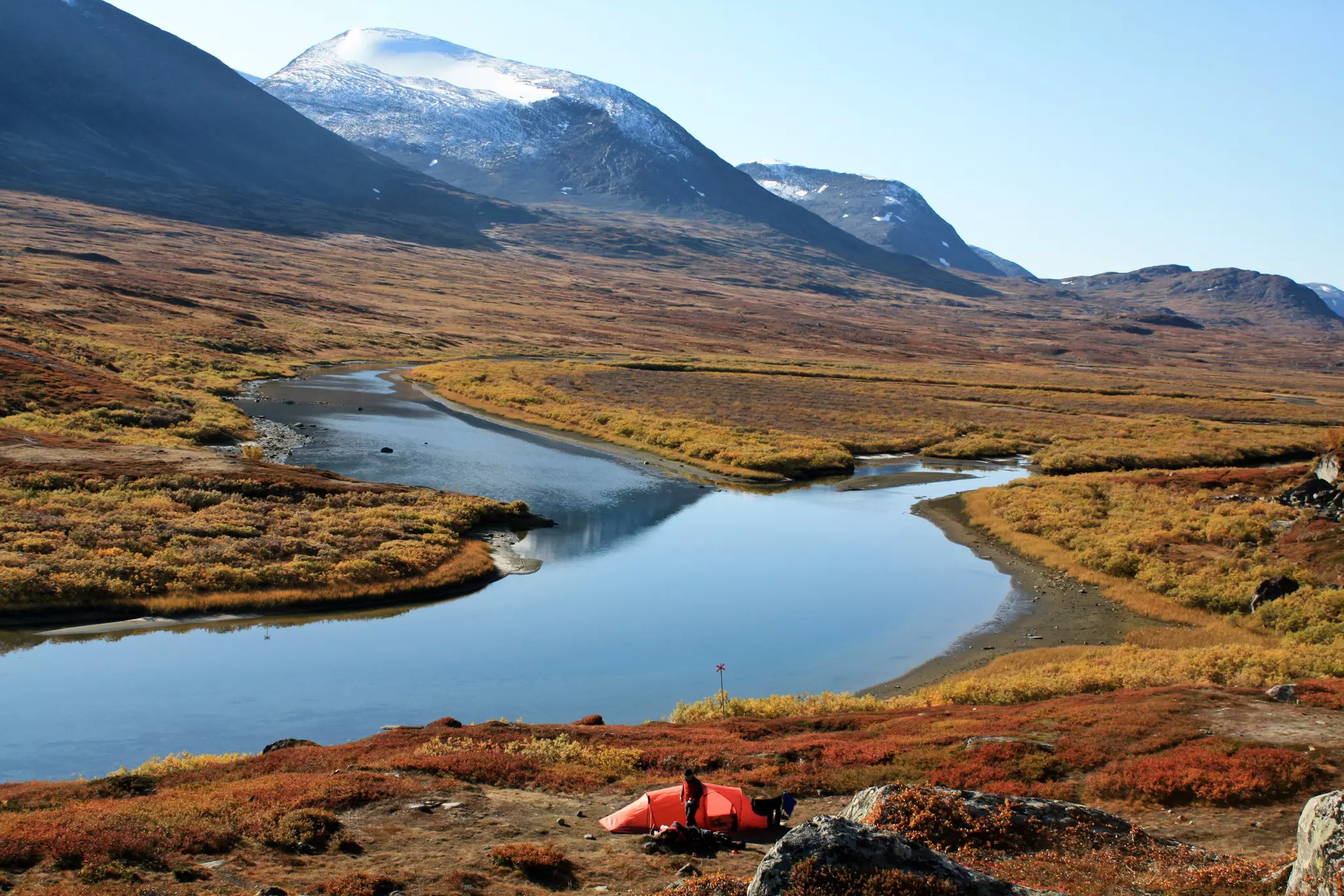 A camper emerges from a small red tent pitched near a river in a mountain valley in summer