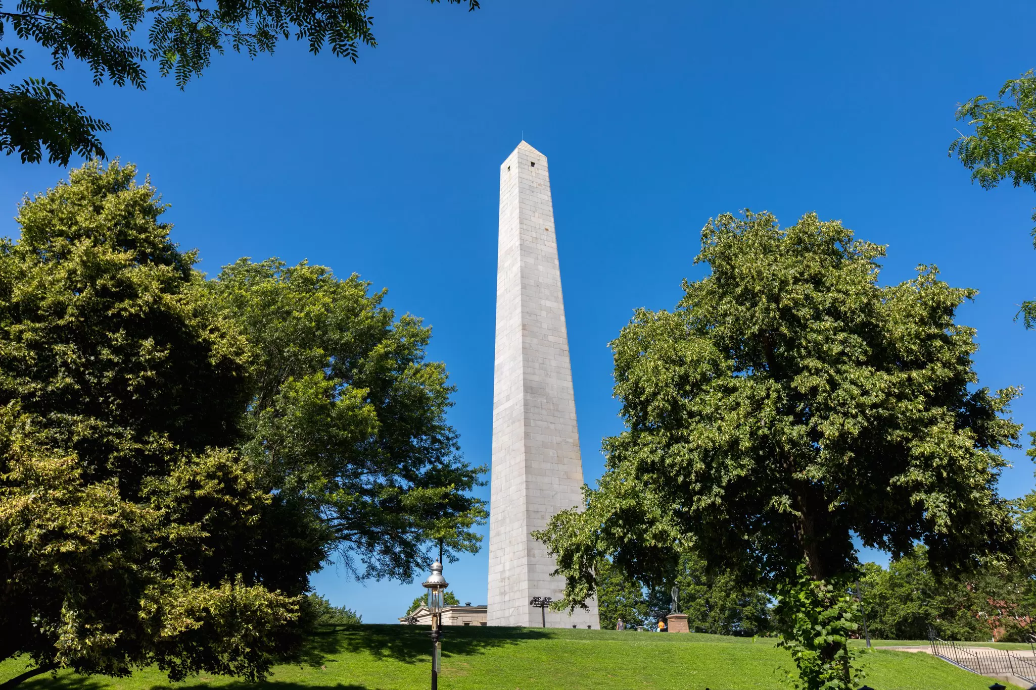 A white obelisk on a green hill under blue sky.