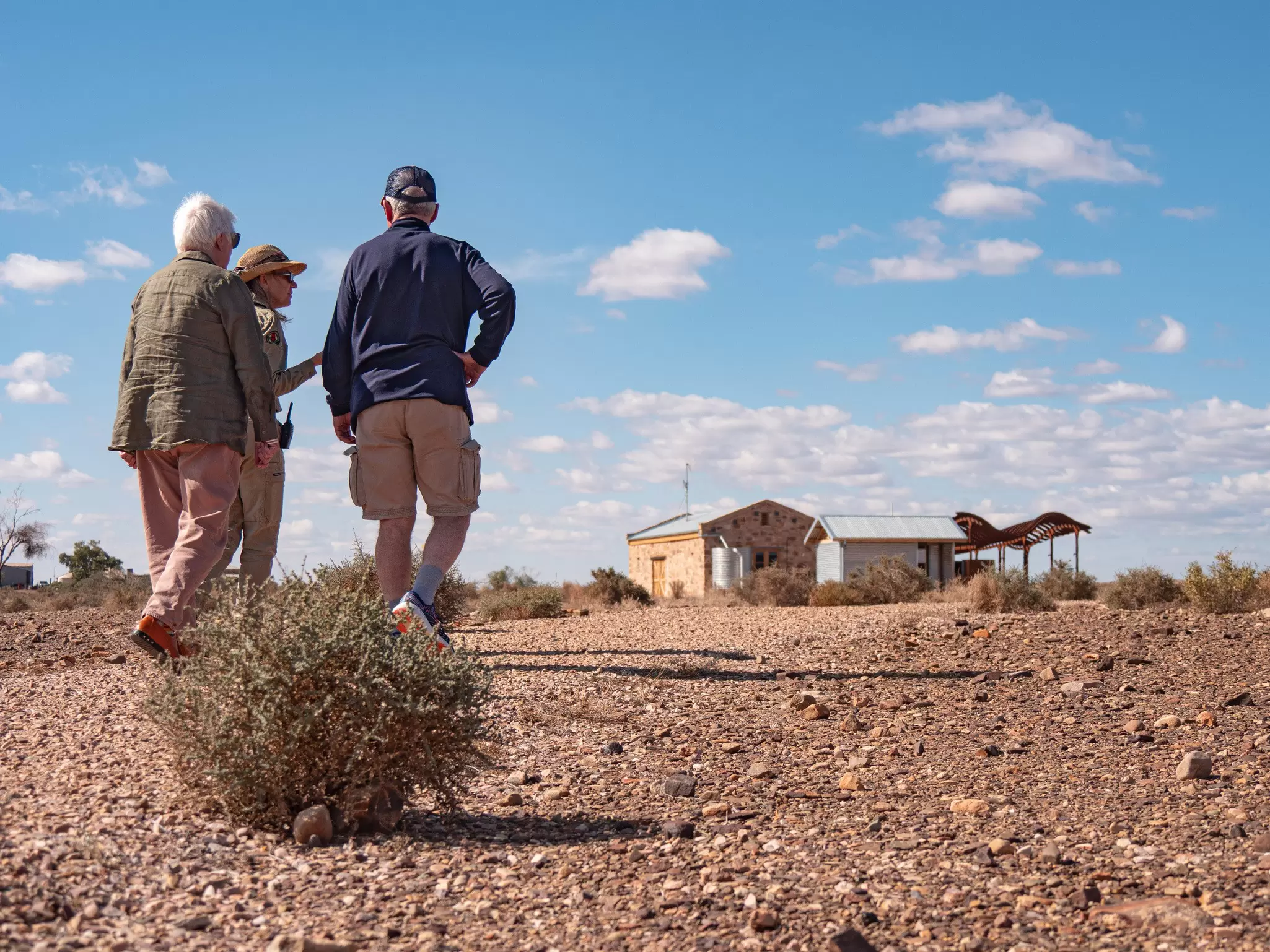 Three people seen from behind walk across stony red outback ground toward a stone and corrugated-iron homestead under a bright blue sky.