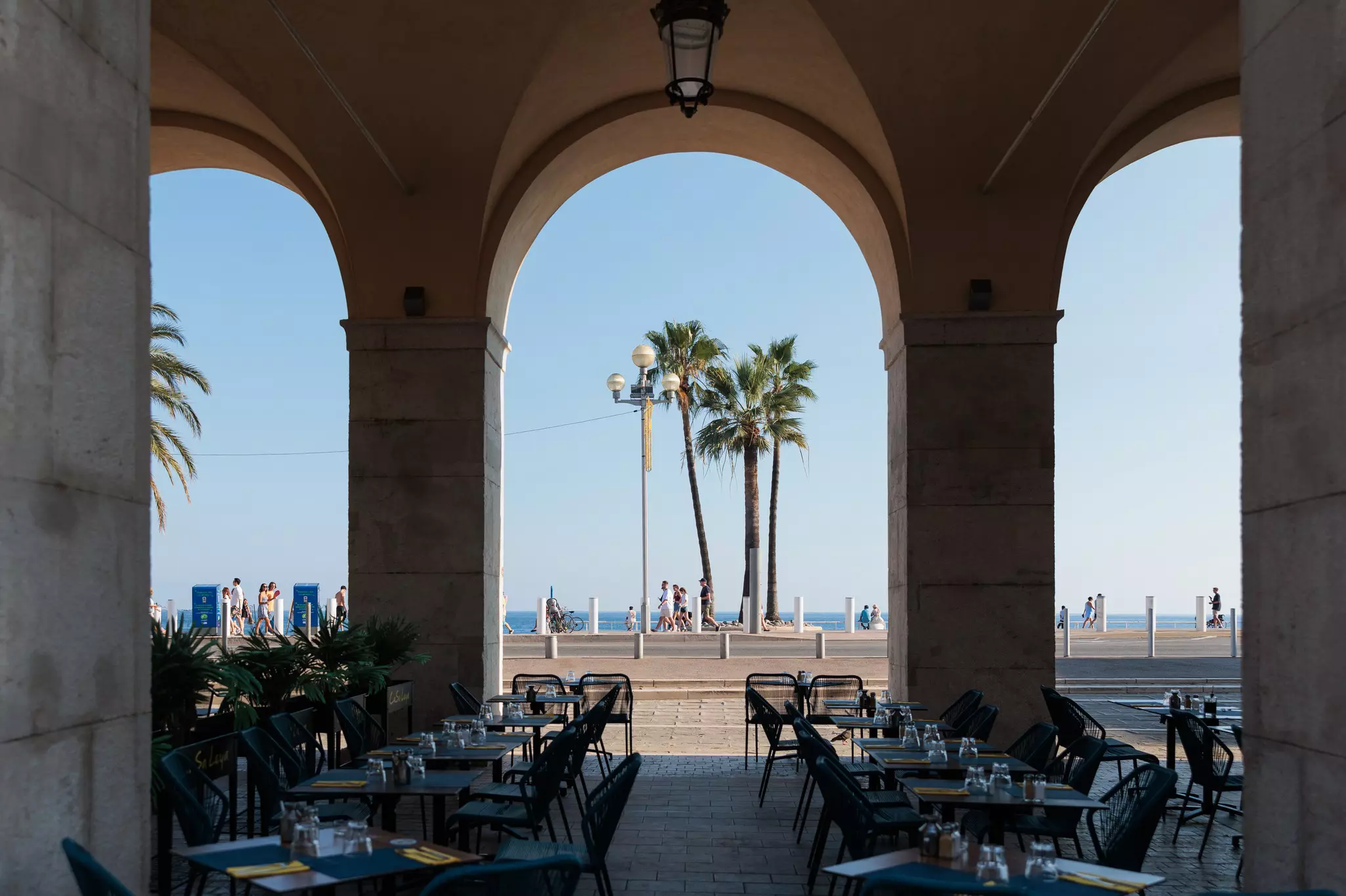 Seating and place settings on a covered terrace. People walking by and a few palm trees are in the background.