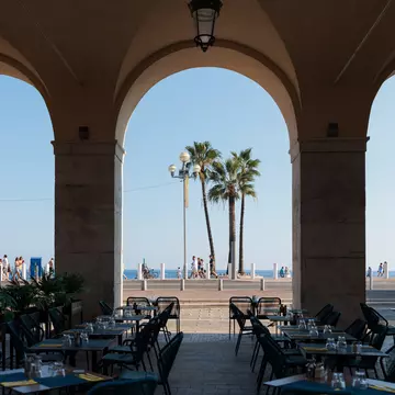 Seating and place settings on a covered terrace. People walking by and a few palm trees are in the background.