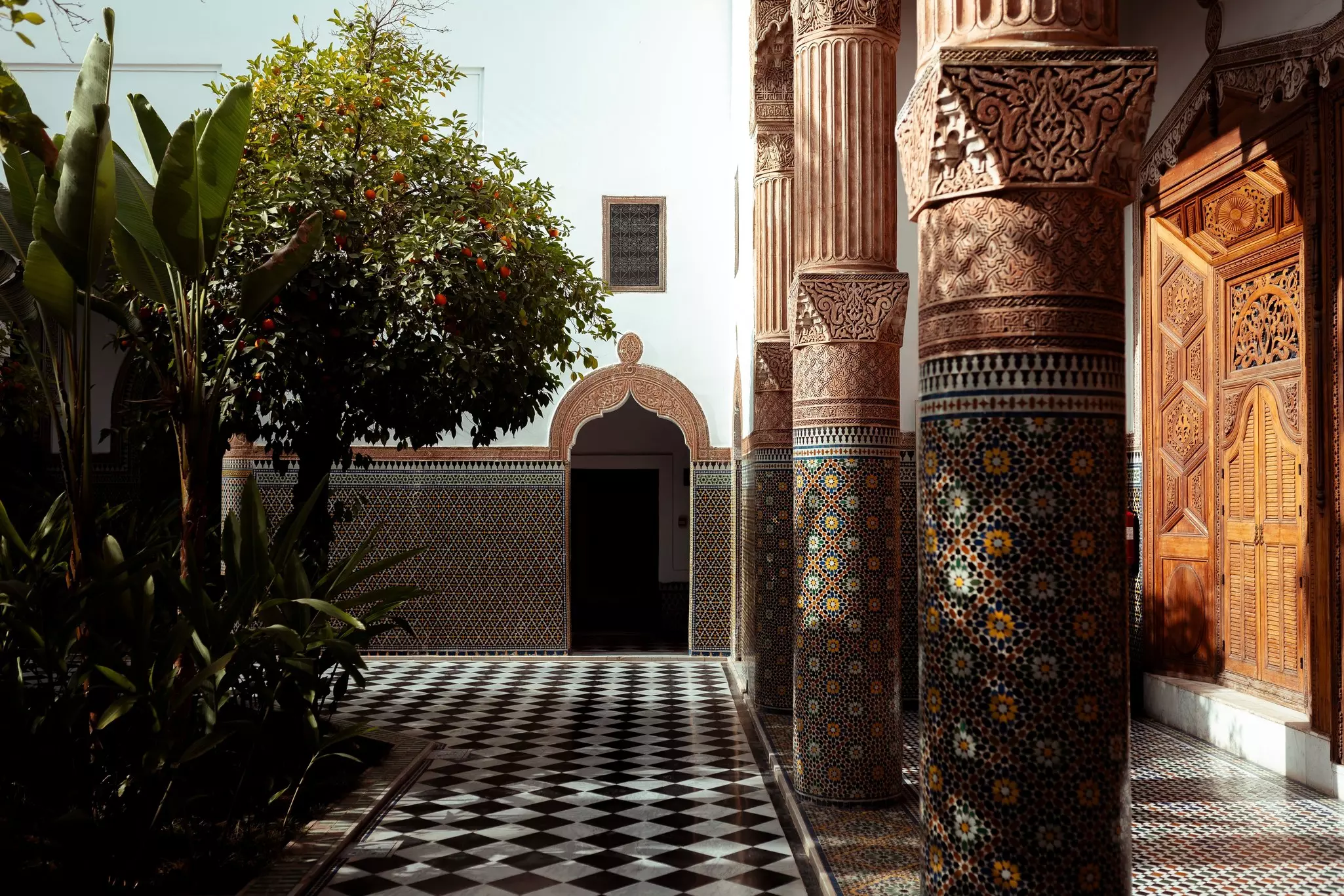 A courtyard with an orange tree, surrounded by intricate tile patterns and carvings on doors, walls, floors and columns.