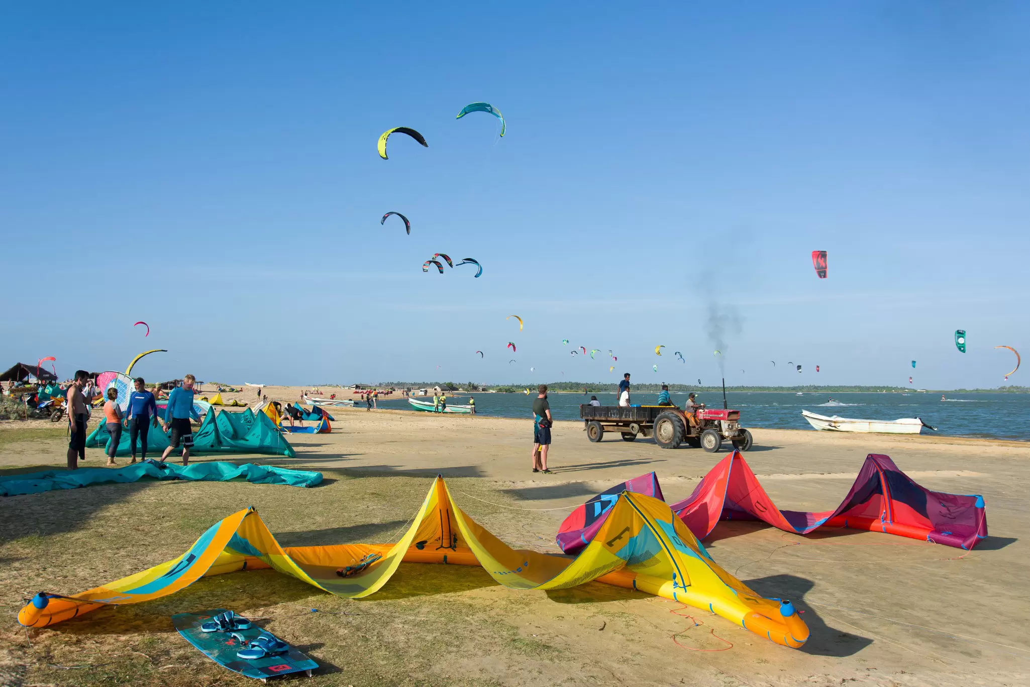 Colorful kites laid out on a beach as people watch kite surfers out on the water