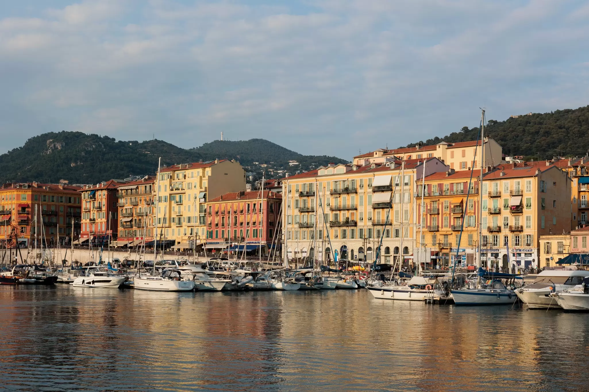Ships in a marina; buildings line the shore a green hill is in the background.