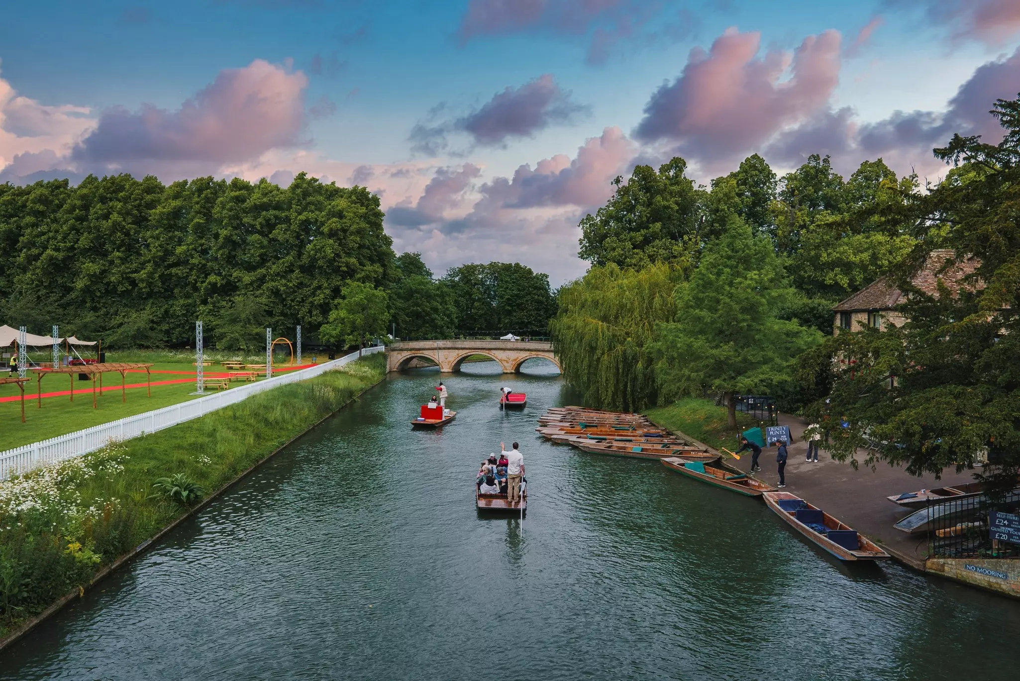 People on rowboats in a canal with a stone bridge and purplish clouds in the distance.