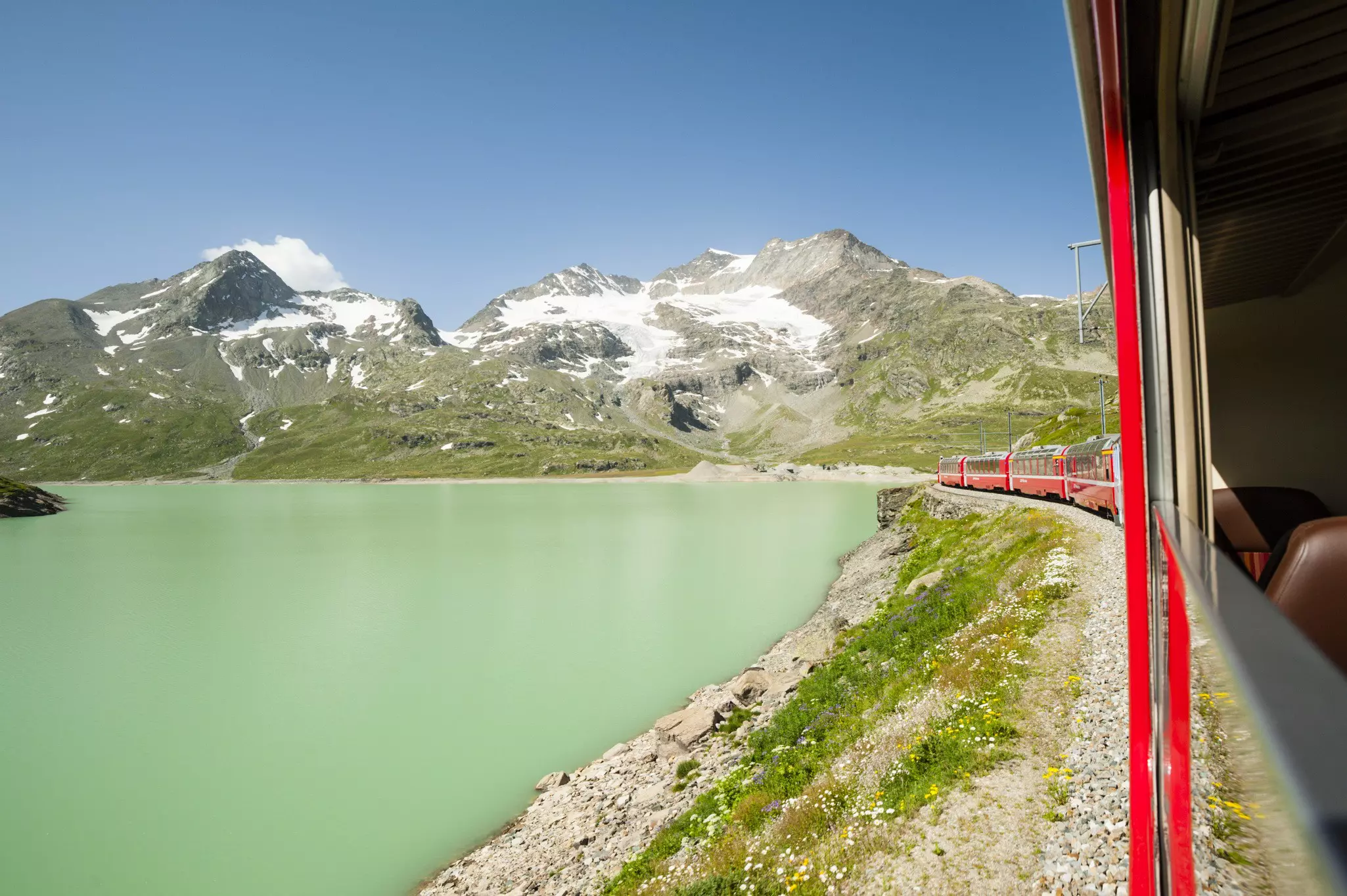 A red train follows a track alongside a green alpine lake heading towards snowy mountain peaks on a sunny day.
