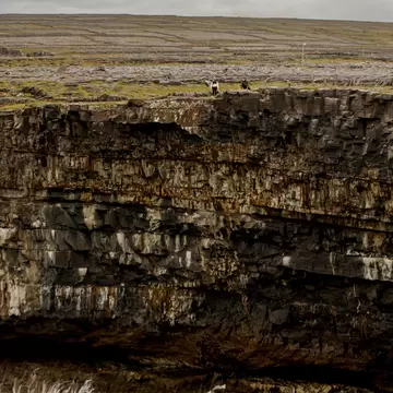 Two people standing on top of tall layered sea cliffs, with waves breaking at the base and flat stony land behind.