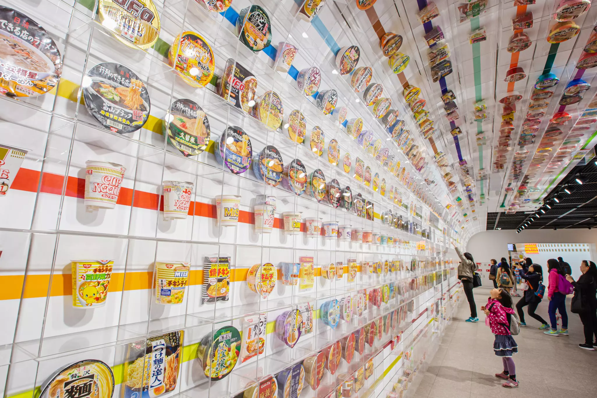 A young girl looks up many cup noodle packaging that decorates a wall in a museum exhibition.