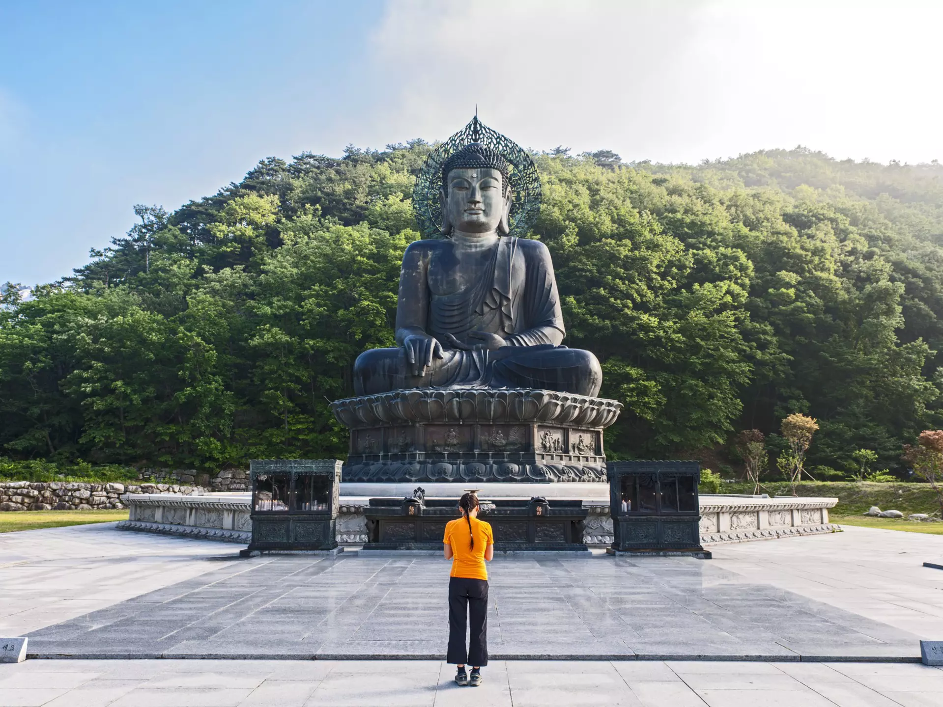 A woman bows her head in front of a large Buddha statue in a rural area surrounded by dense woodland.