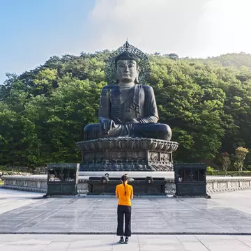 A woman bows her head in front of a large Buddha statue in a rural area surrounded by dense woodland.