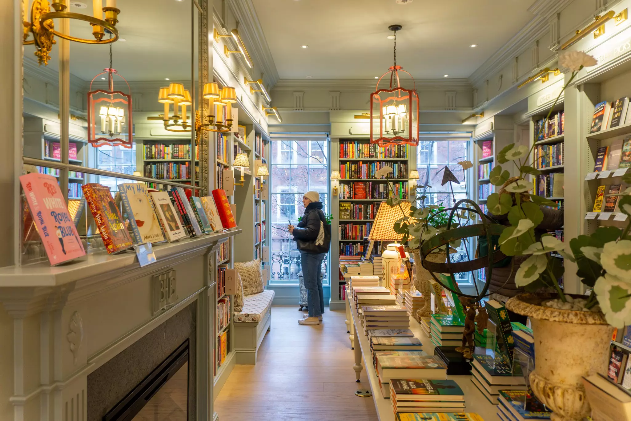 A person dressed in winter clothes pauses to browse books that line the shelves of a cozy store.