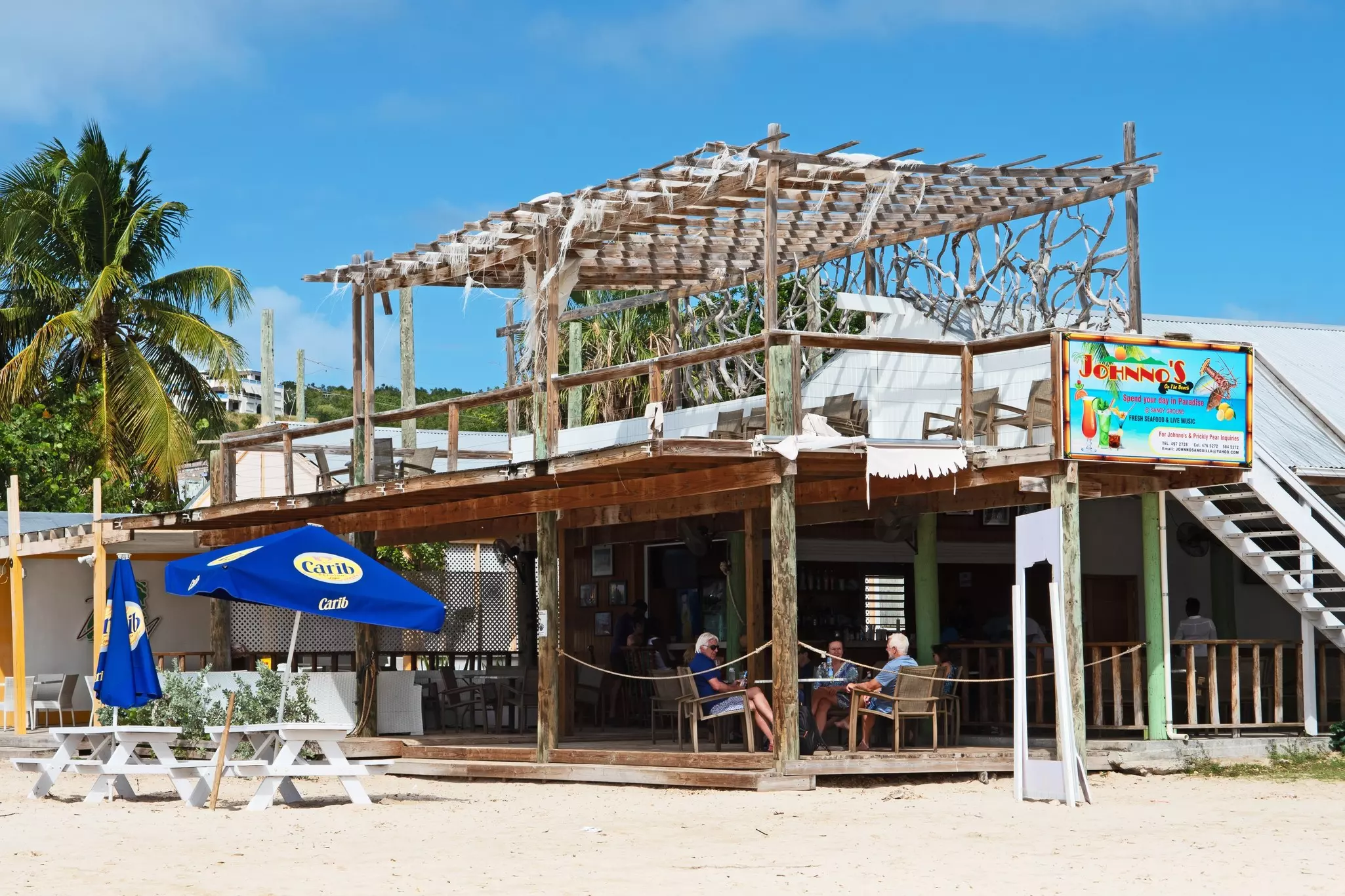 A beachfront bar and restaurant. White picnic tables with blue umbrellas are set on the sand. Three people sit on the wooden restaurant's terrace.