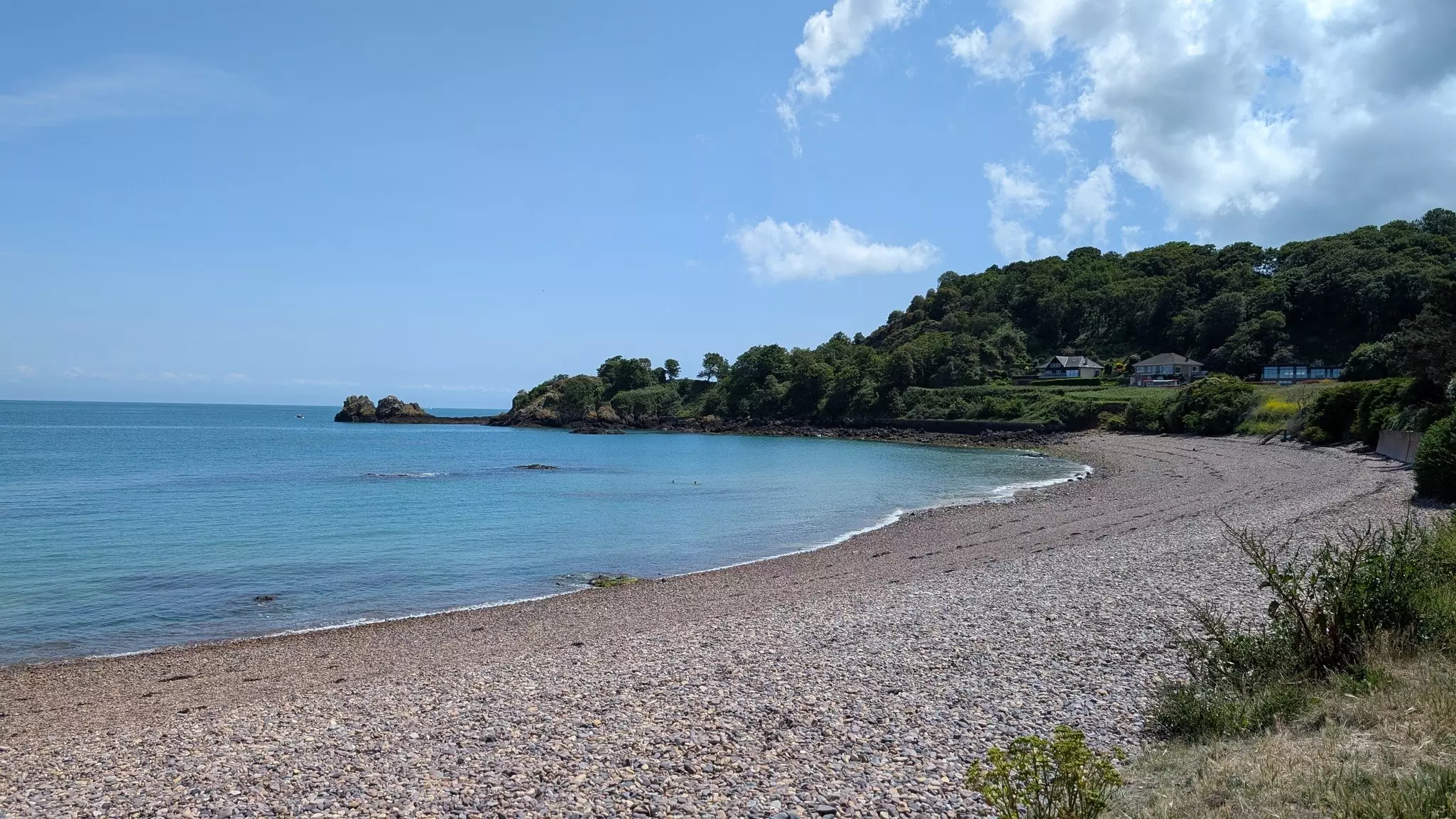 Rocky curved beach with trees and homes in the distance and the ocean to the left on a mostly sunny day.