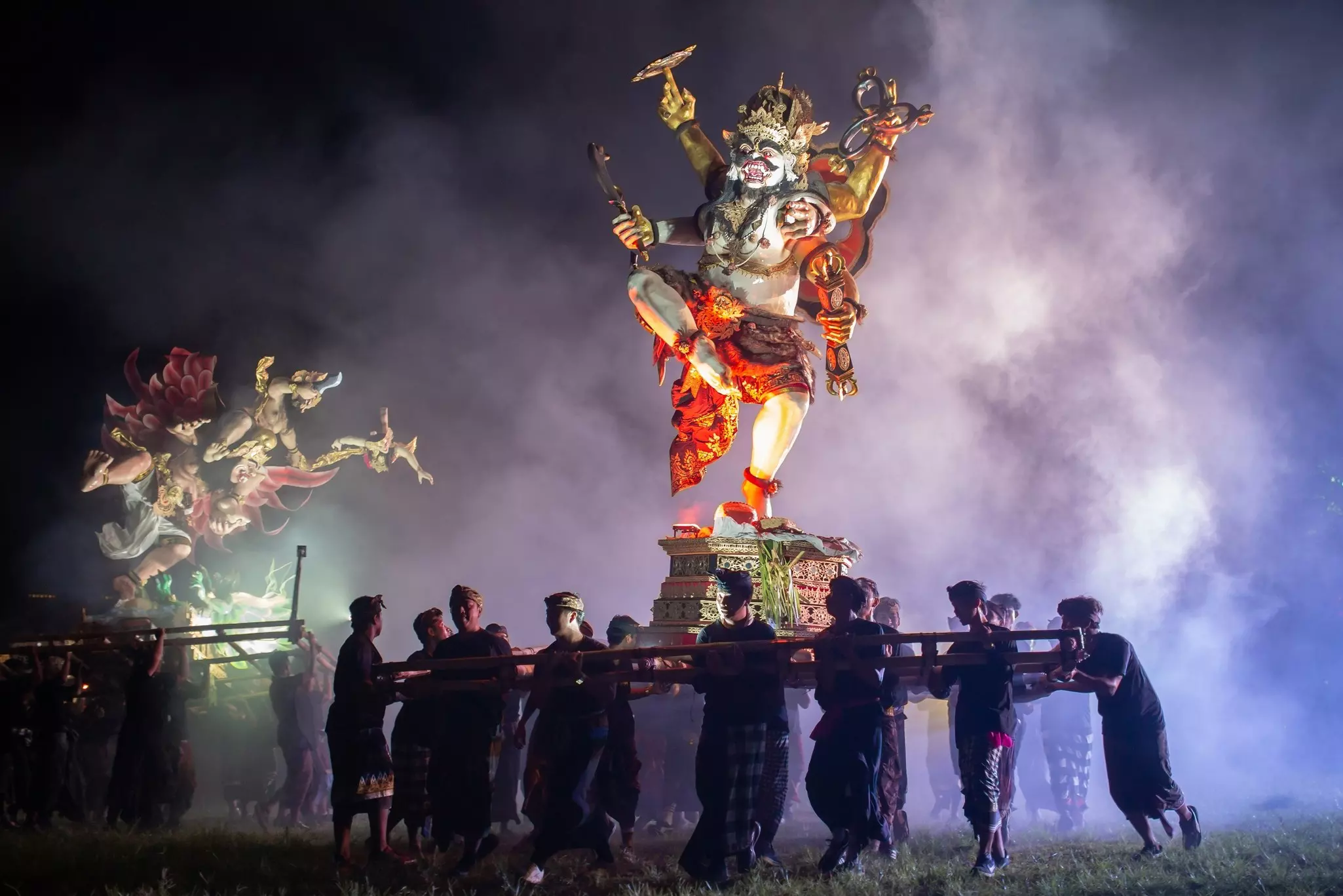 A large statue of a demon carried by a group of people with smoke in the background at night.