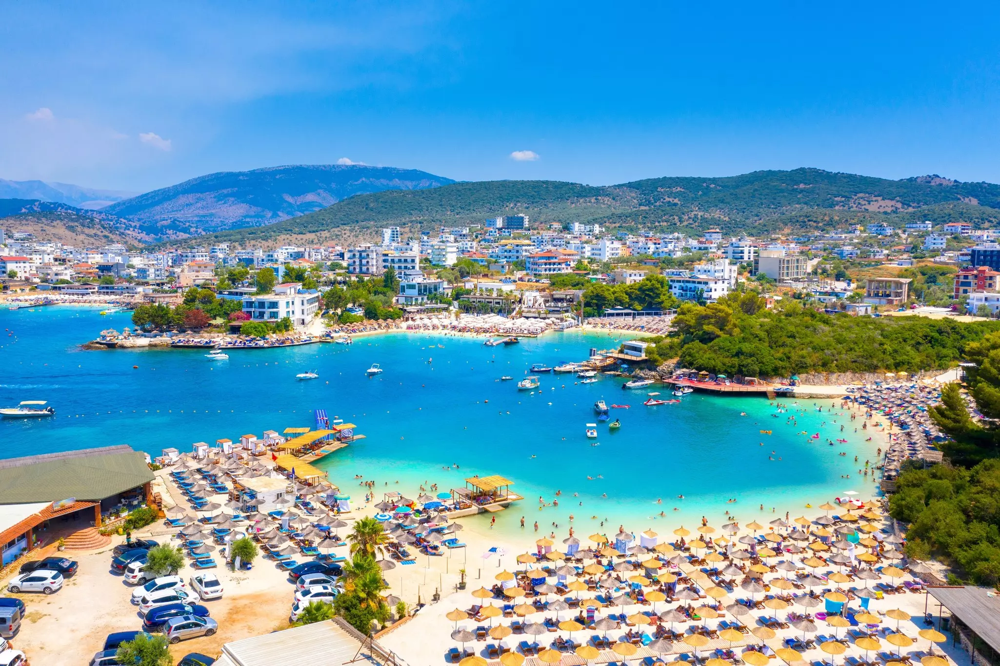 Bora Bora beach in Ksamil, Albanian Riviera, with cars parked on one side and umbrella-topped lounging stations on another