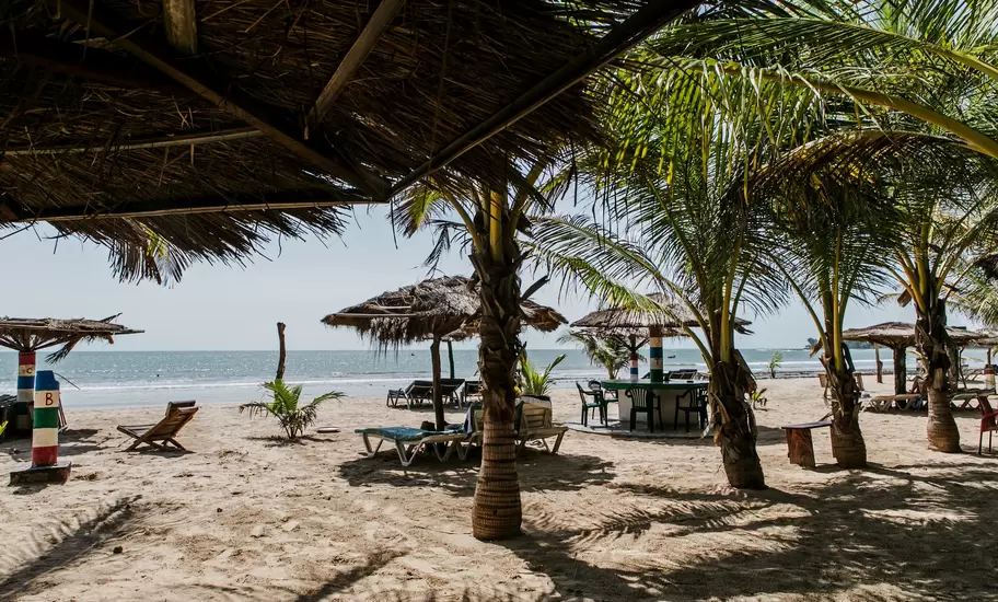 Beach bar in The Gambia with thatched shelters, palms, loungers and posts painted in red, blue and green bands, sea beyond