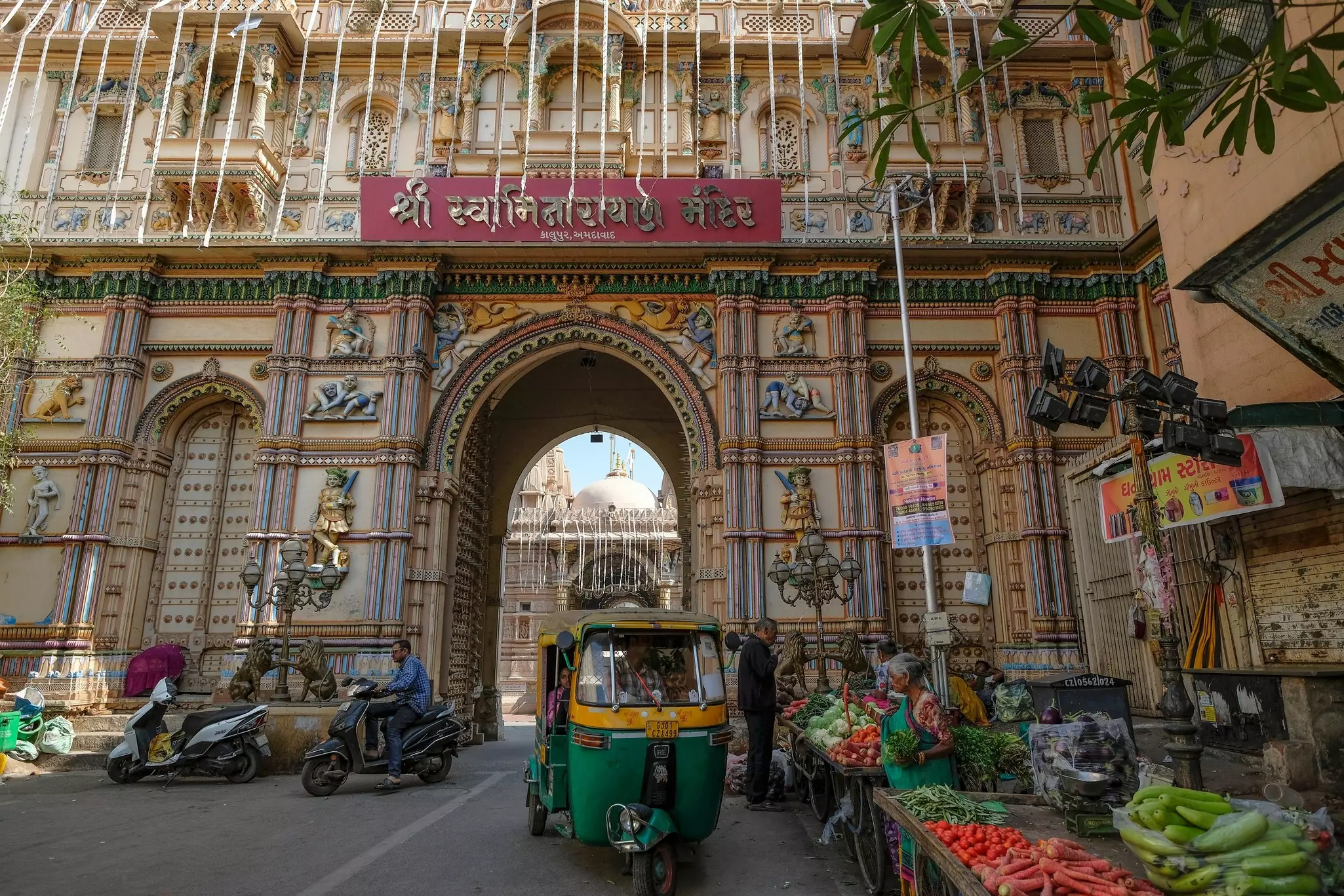 A tuk tuk drives past vegetable vendors. Behind it is an arch.