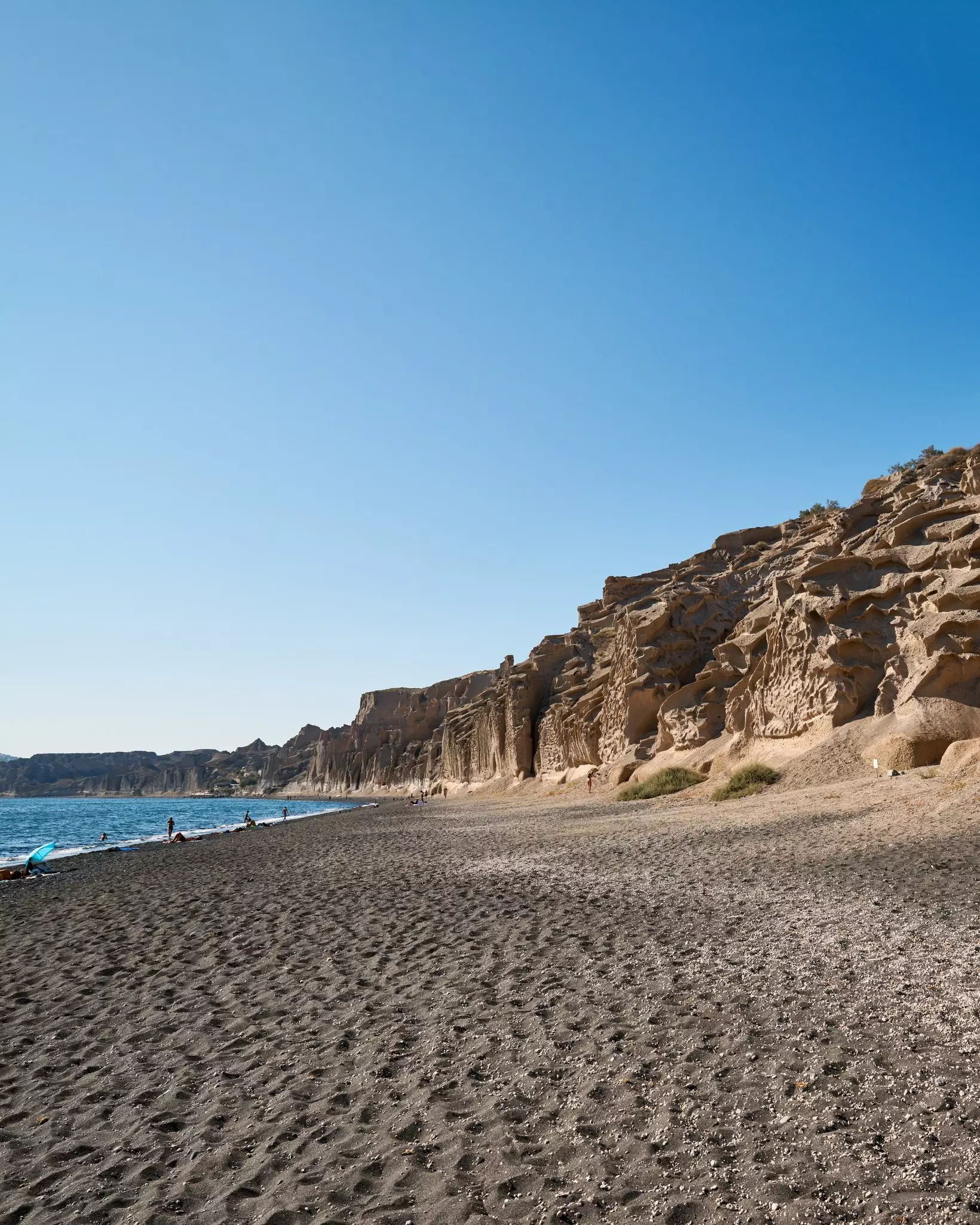 Beach with high, craggy cliffs rising from the sand. Beachgoers linger near the shore.