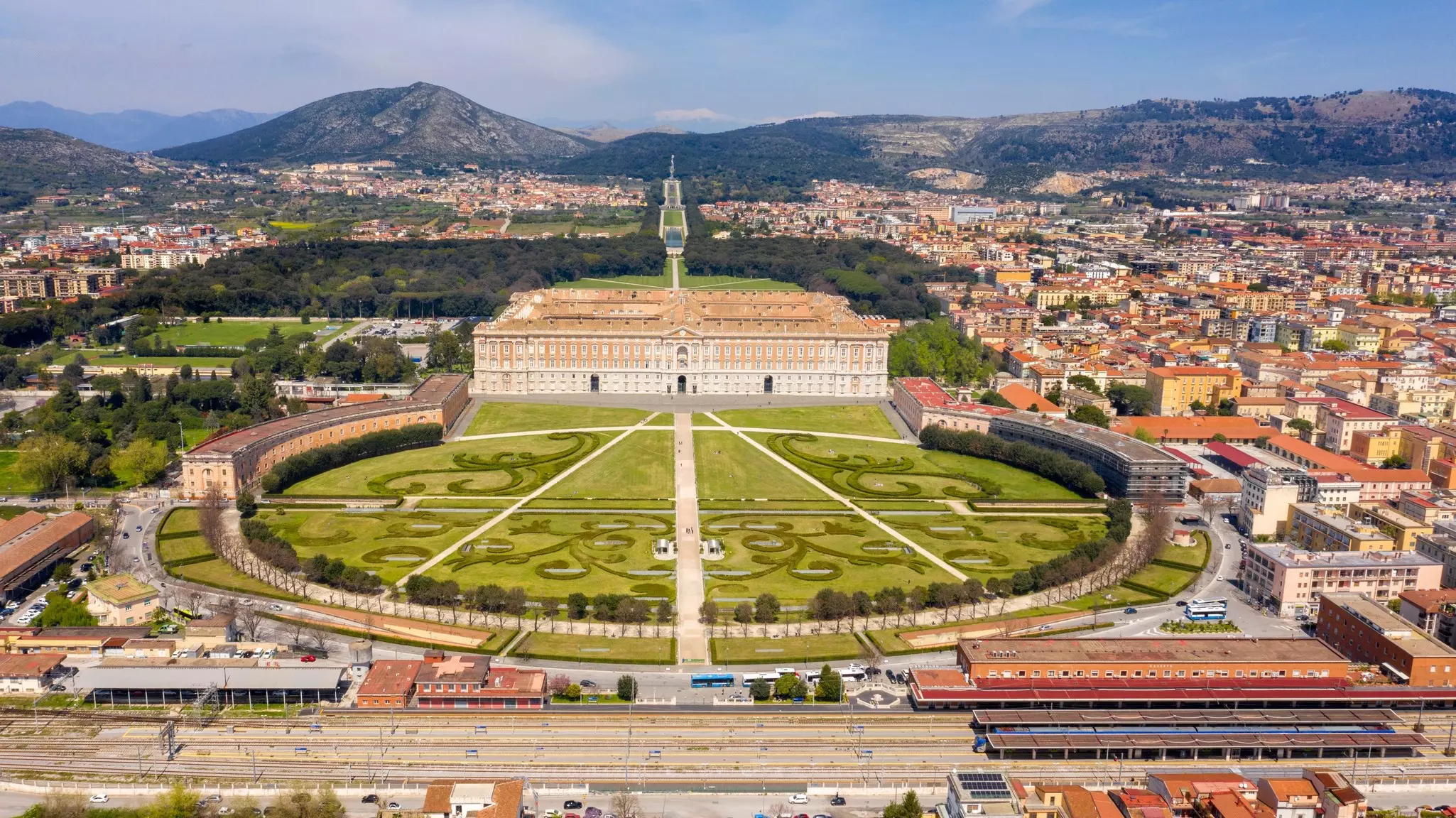 An aerial view of Reggia di Caserta in Campania near Naples, a large palace with a round garden complex containing decorative shrubbery and paths leading to the building's entrance. Residential buildings and mountains can be seen in the background.