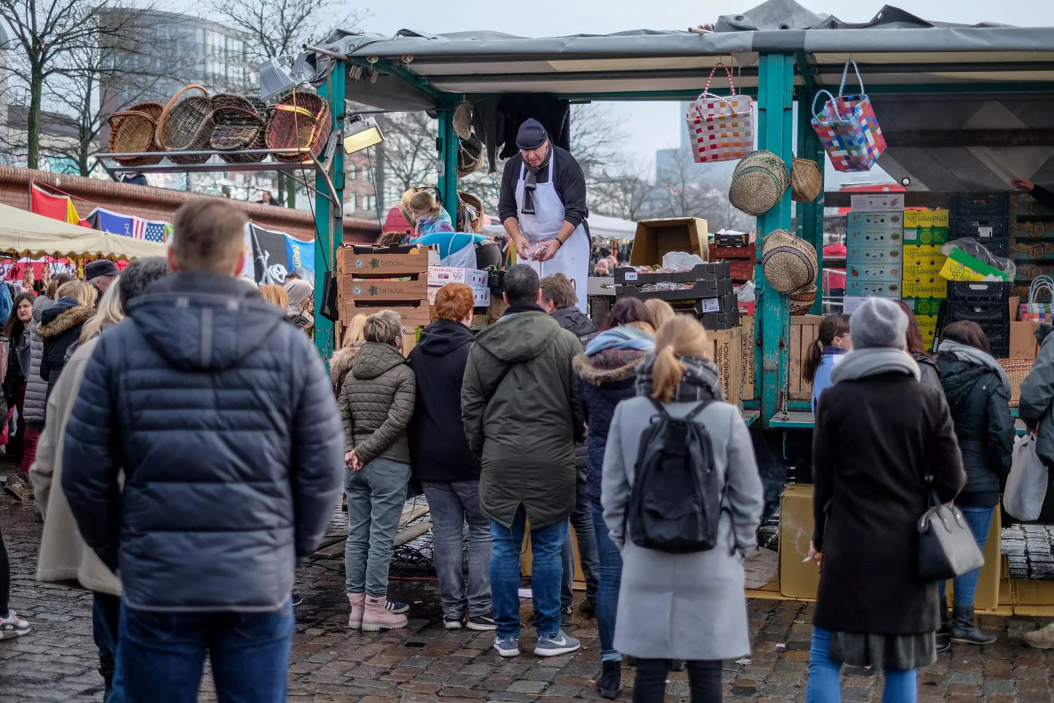 A crowd of people in winter coats waits at an outdoor vendor's busy stall on an overcast day.