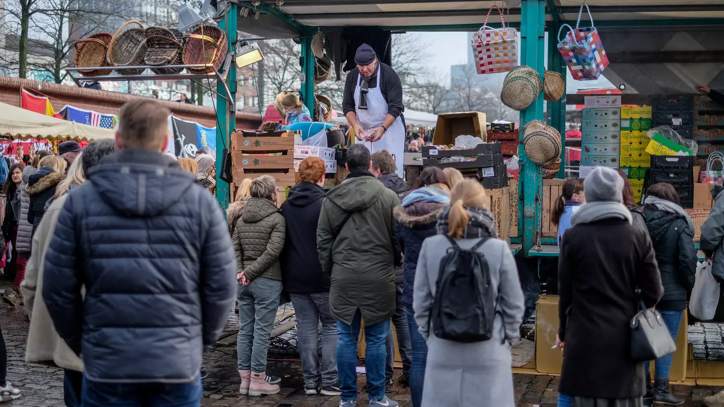 A crowd of people in winter coats waits at an outdoor vendor's busy stall on an overcast day.