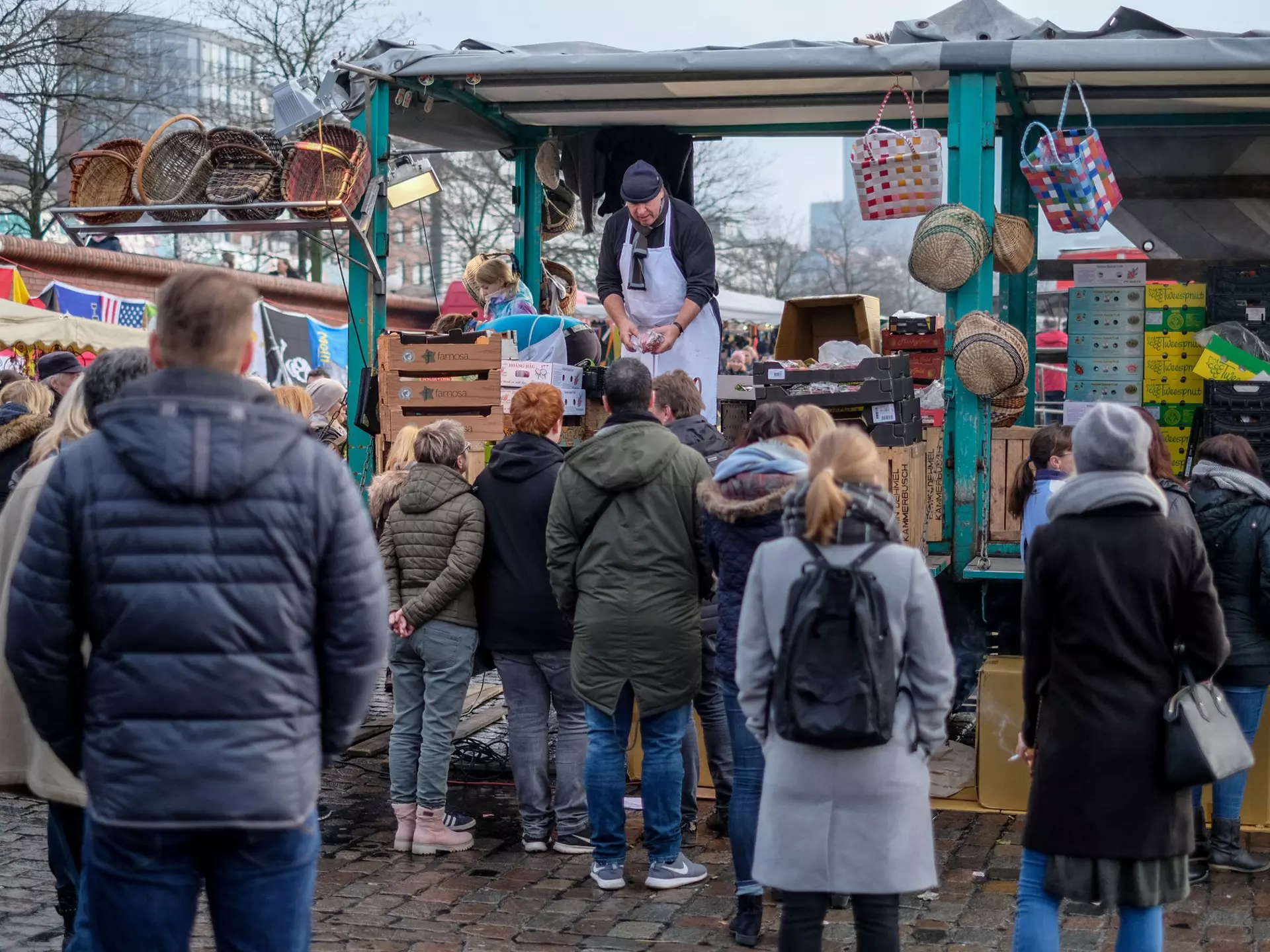 A crowd of people in winter coats waits at an outdoor vendor's busy stall on an overcast day.