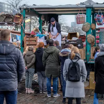 A crowd of people in winter coats waits at an outdoor vendor's busy stall on an overcast day.