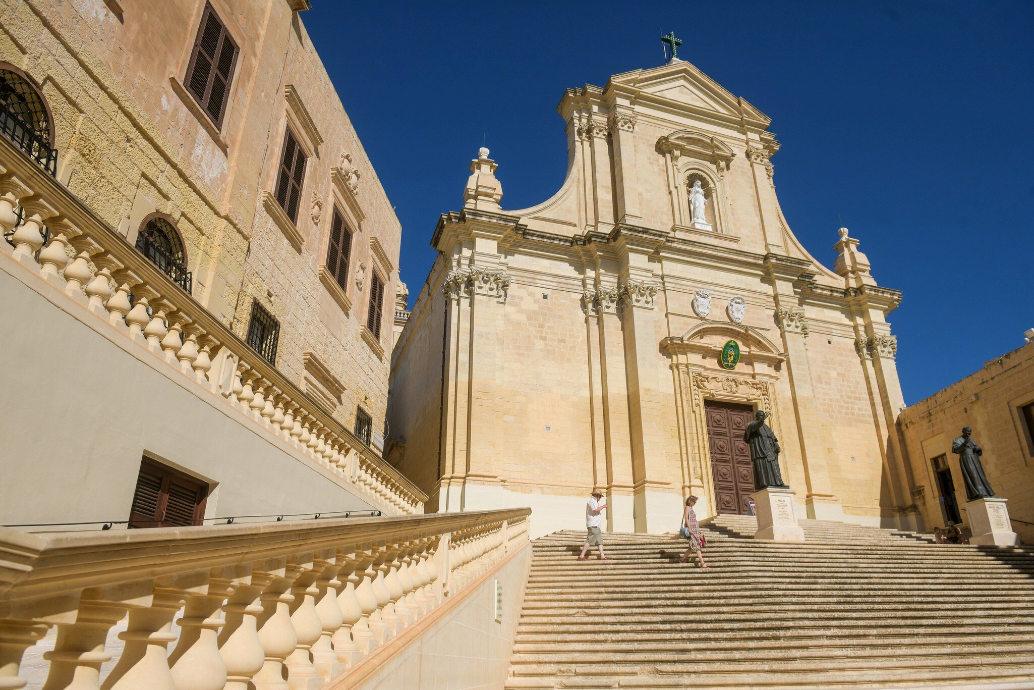 Baroque limestone cathedral facade at the top of wide stone steps in Gozo's Cittadella, with two people climbing.