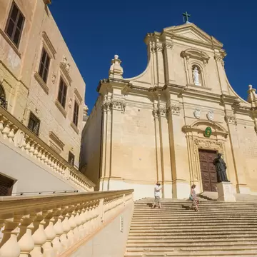 Baroque limestone cathedral facade at the top of wide stone steps in Gozo's Cittadella, with two people climbing.