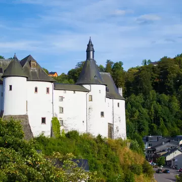 Château de Clervaux, a whitewashed castle with dark conical towers on a wooded slope above houses in Clervaux