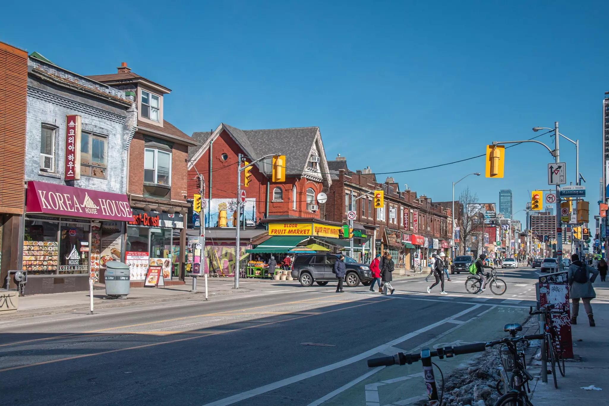 Busy intersection on Bloor Street West in Toronto’s Koreatown, with diverse shops, cyclists, and pedestrians under a clear blue sky.