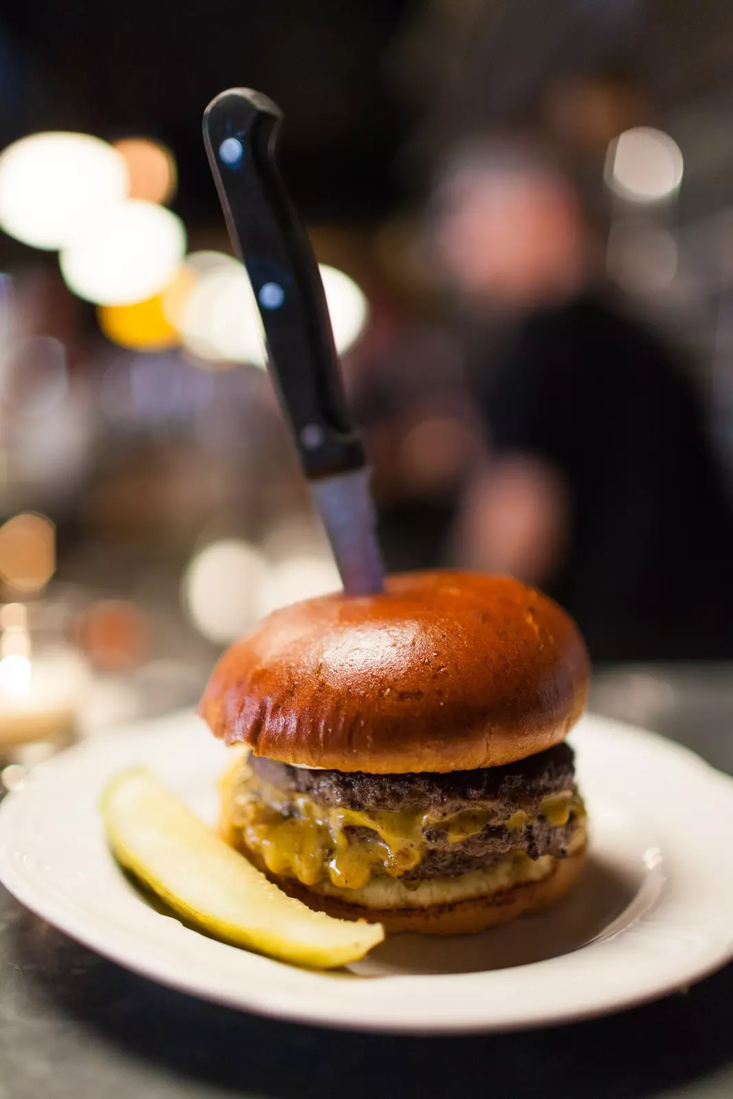A double cheeseburger with a pickle on the side and a knife stuck on top.