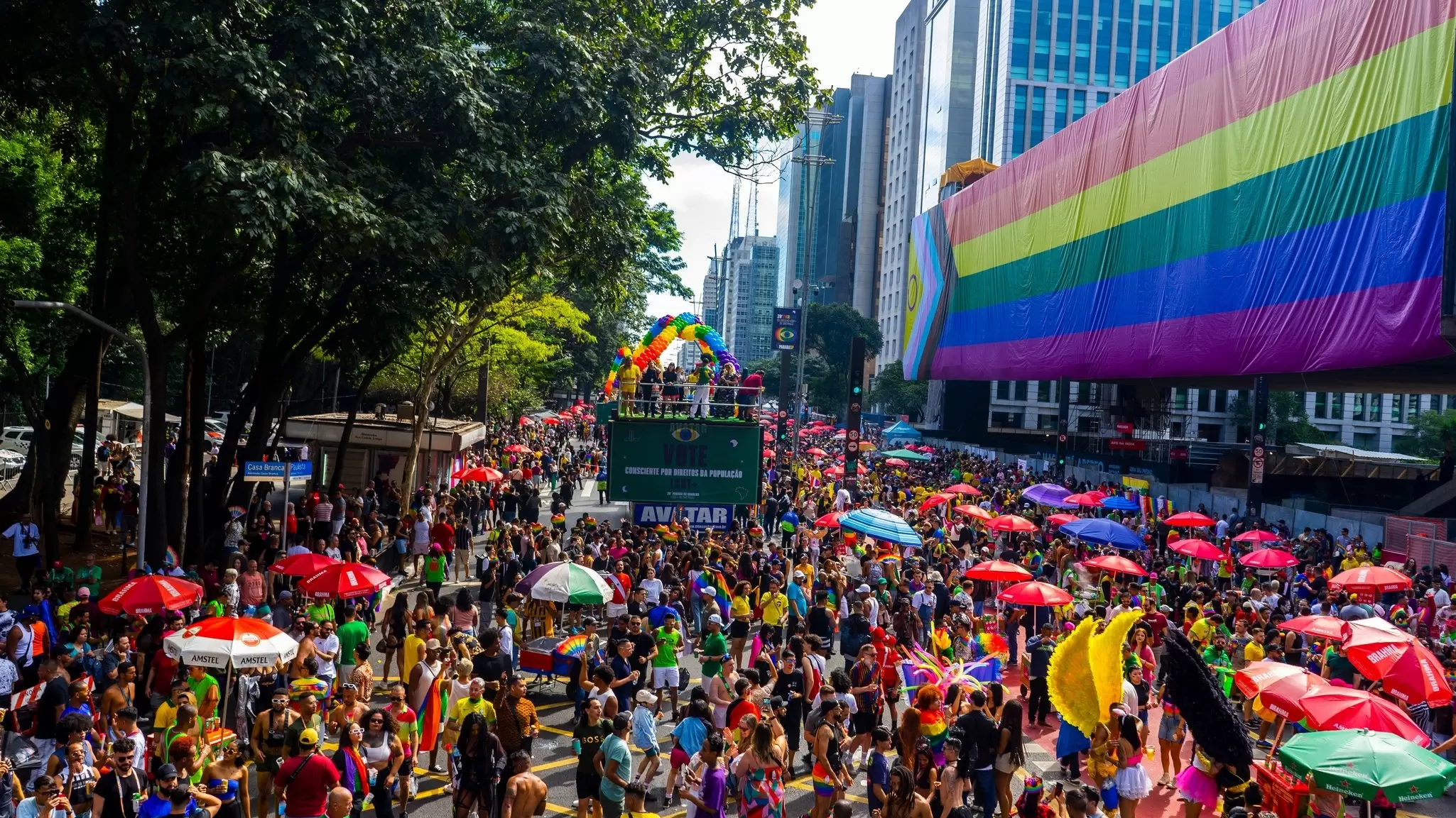 People fill a street, some with sun umbrellas; a rainbow flag is displayed on a building to one side, and a rainbow balloon arch is in the distance.