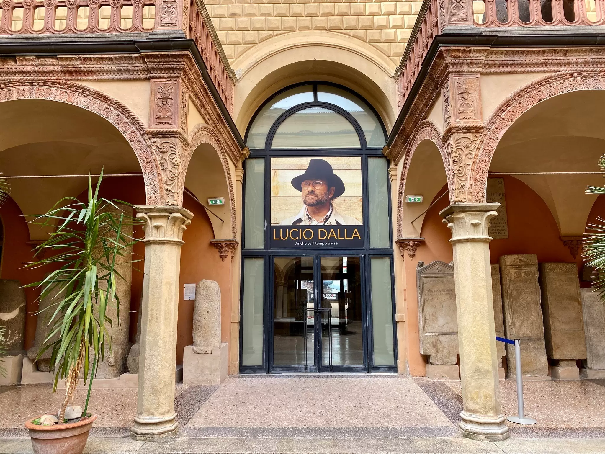 An arched museum entrance in a Bologna courtyard displays a large Lucio Dalla exhibition poster.