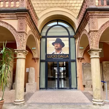 An arched museum entrance in a Bologna courtyard displays a large Lucio Dalla exhibition poster.
