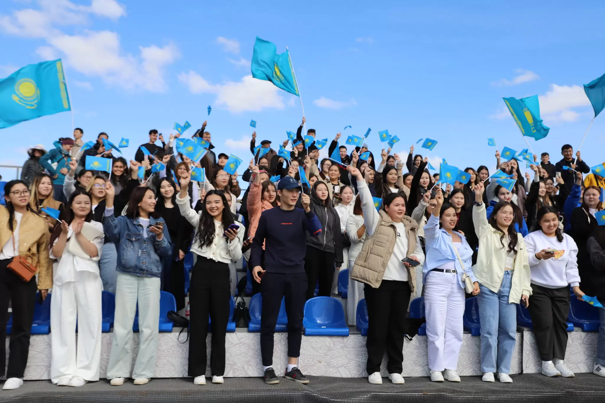 Groups of spectators cheer and wave Kazakhstan's blue and yellow national flag on the side of the game arena at the World Nomad Games in 2024 